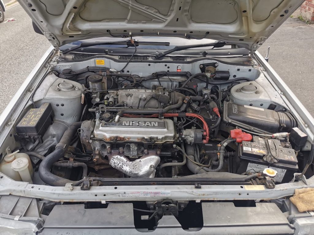 Engine bay of the Nissan Bluebird showing the Nissan-branded cam cover, with various hoses, wiring, and ancillaries visible.
