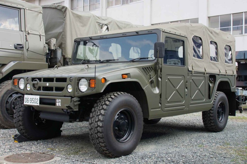 A Japanese Ground Self-Defence Force High Mobility Vehicle, finished in olive drab with a canvas soft top and large all-terrain tyres, parked at a military base.