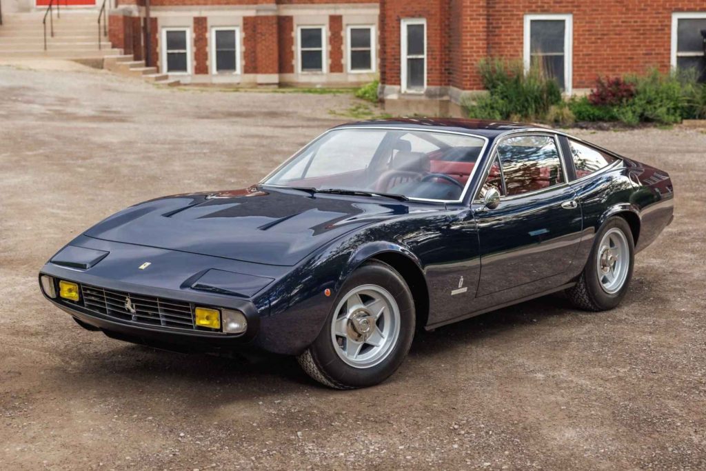 1971 Ferrari 365 GTC/4 in dark navy blue, three-quarter front view, parked in front of a red brick building with yellow fog lights and five-spoke alloy wheels