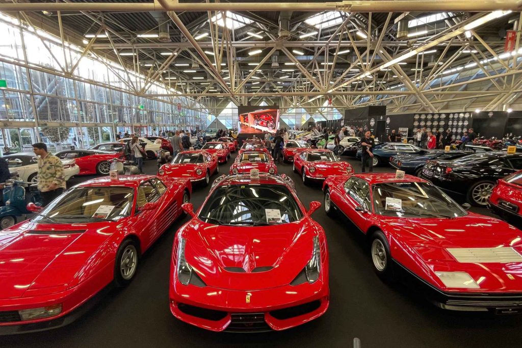 An indoor automotive show floor packed with red Ferraris including a Testarossa, 488, and Dino, with spectators browsing between them.