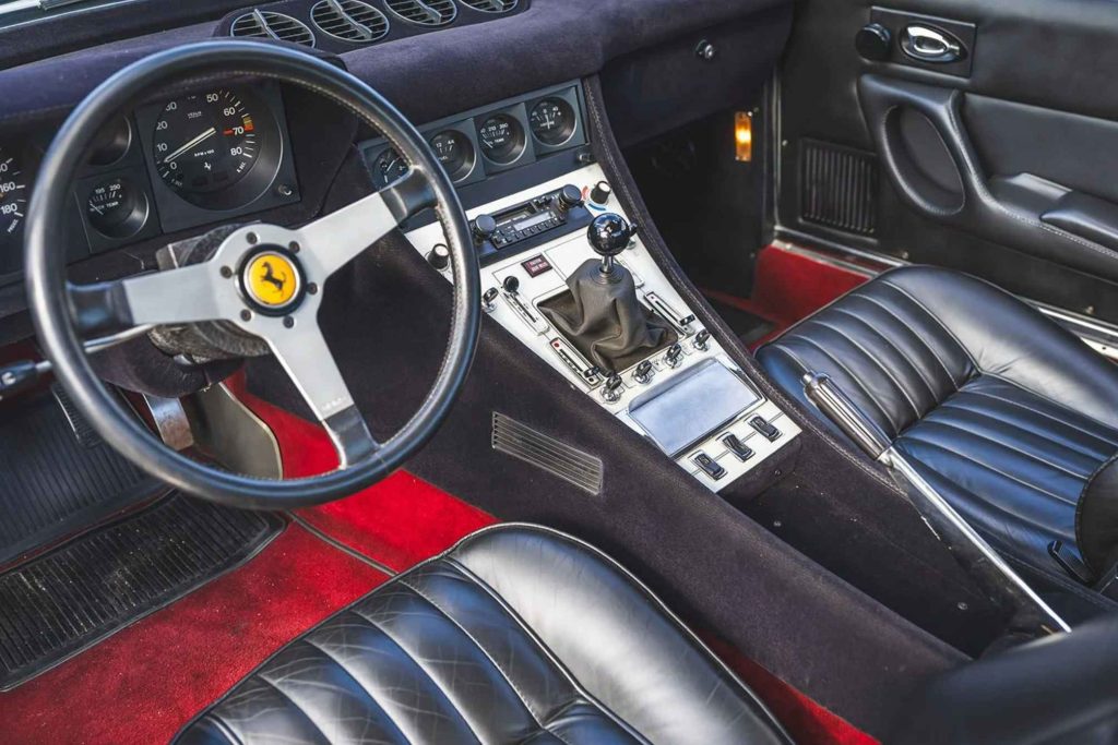 Interior of a Ferrari 365 GTC/4 showing the black leather seats, red carpet, three-spoke Momo steering wheel with prancing horse badge, center console with gear shifter, and analog instrument cluster