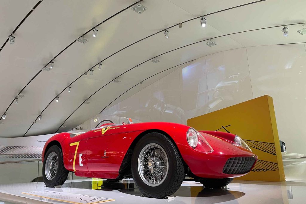 A red vintage Ferrari racing spider, numbered 7 and bearing "Scuderia Ferrarano" livery, displayed in a curved white museum gallery.