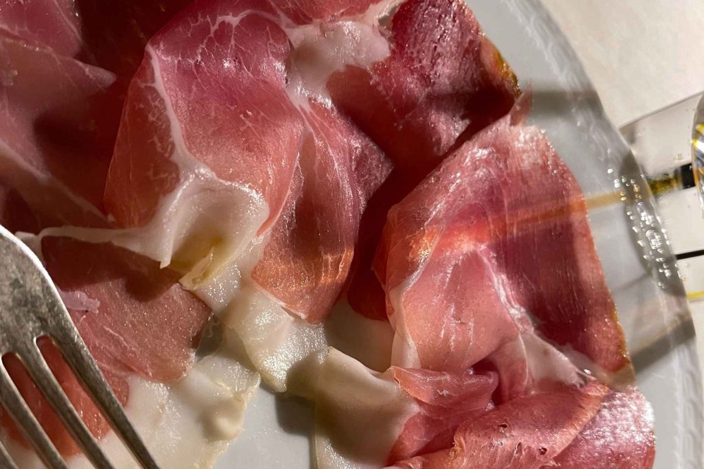 Thinly sliced Italian cured meat, likely culatello or prosciutto, draped on a white plate with a fork visible at the edge.