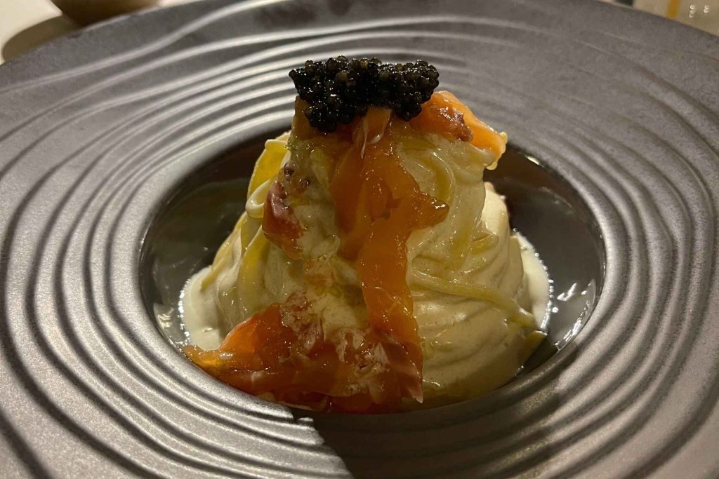 A mound of delicate pasta topped with salmon and black caviar, served in a ridged gray ceramic bowl.