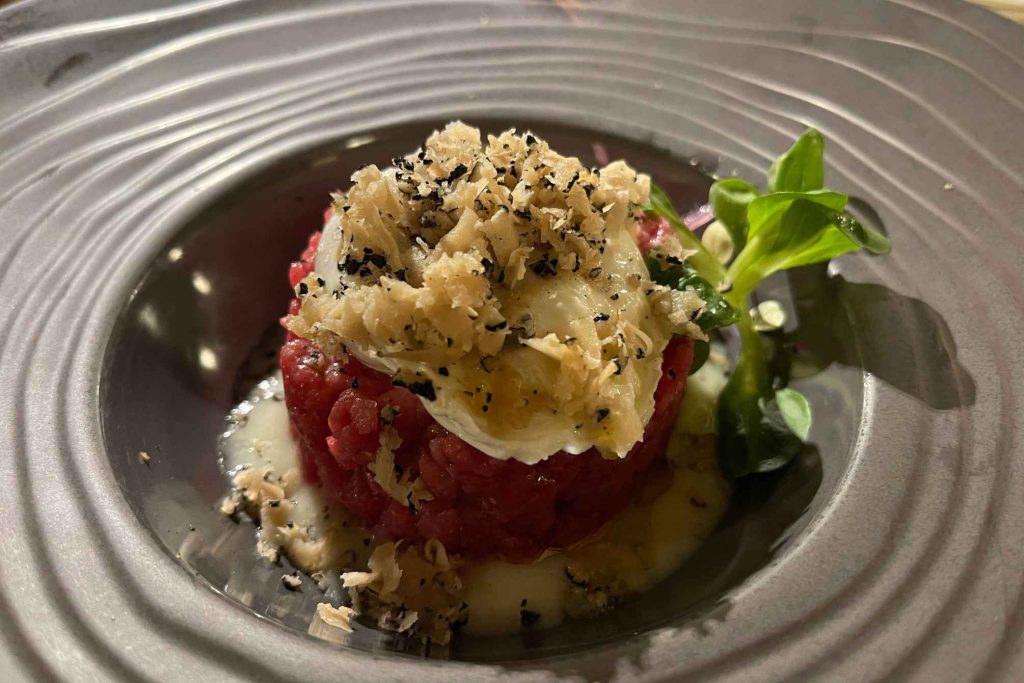 A fine-dining beef tartare tower topped with shaved aged cheese and black pepper, served with a small green salad garnish in a ridged grey bowl.
