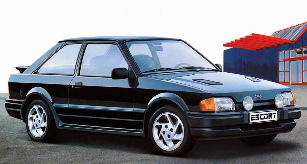 A black Ford Escort RS Turbo three-door hatchback photographed in a three-quarter front view against a white background, with a colorful industrial building in the background featuring red and blue panels.