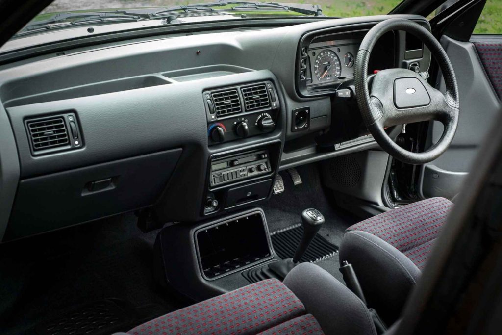 The right-hand-drive interior of a Ford Escort RS Turbo, showing the grey dashboard, cassette head unit, manual gearshift, and grey and red patterned sports seats.