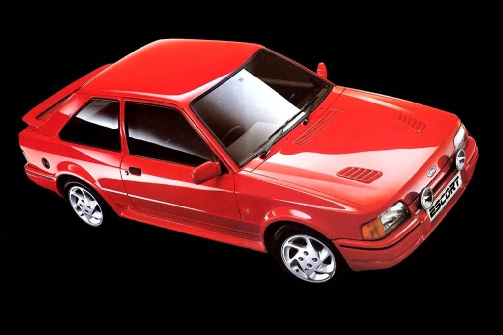 A red Ford Escort RS Turbo photographed from a high three-quarter front angle against a black background, showing the bonnet vents, wide body kit, and five-spoke alloy wheels.