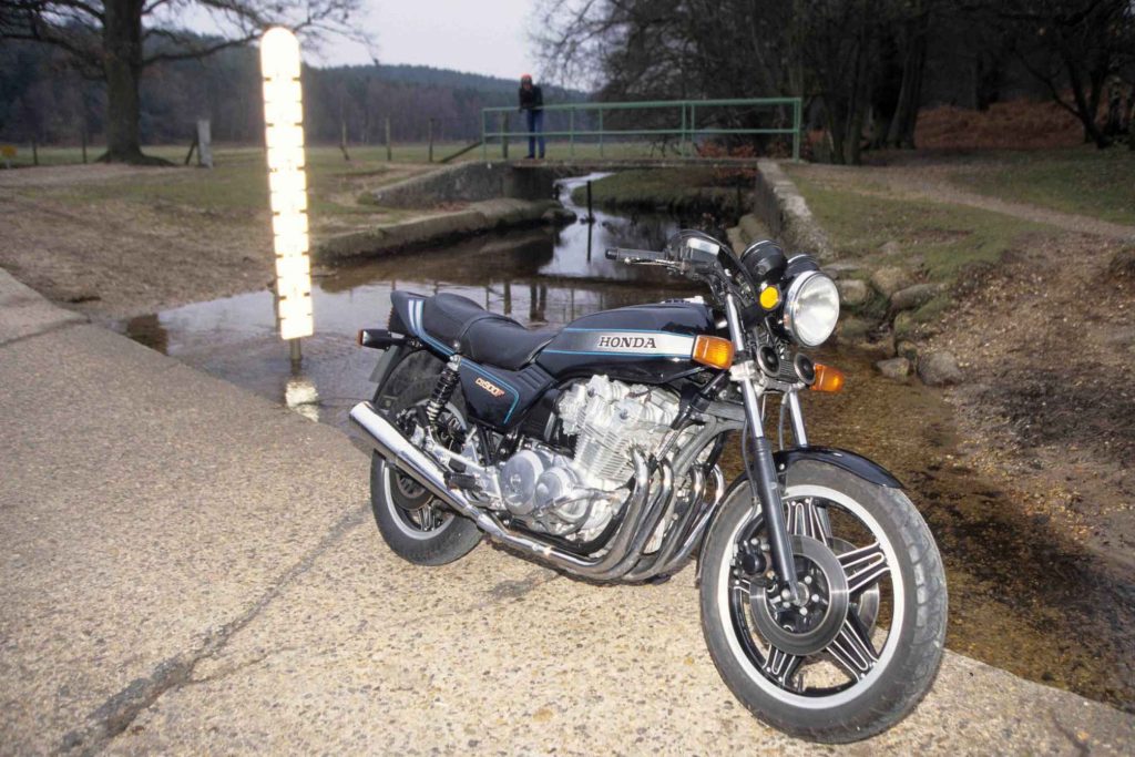 A Honda CB900F motorcycle parked beside a narrow concrete-banked canal in a rural park setting, shot at dusk with a glowing marker post nearby and a person standing on a bridge in the background.