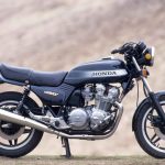 Right-side profile studio-style shot of a Honda CB900F motorcycle in black with silver and blue tank graphics, displayed on its center stand against a sandy hillside backdrop.