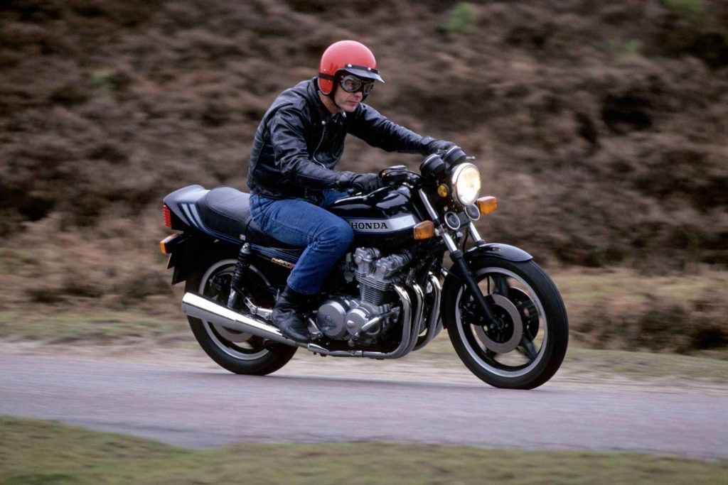 A rider in a red open-face helmet, goggles, and black leather jacket rides a Honda CB900F at speed on a country road, background blurred with motion.