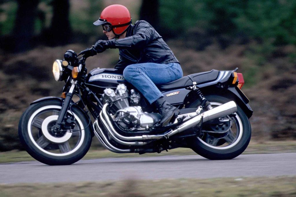 Rear three-quarter action shot of a Honda CB900F with the rider leaning forward, showcasing the four-into-four chrome exhaust headers and whitewall-rimmed rear wheel.
