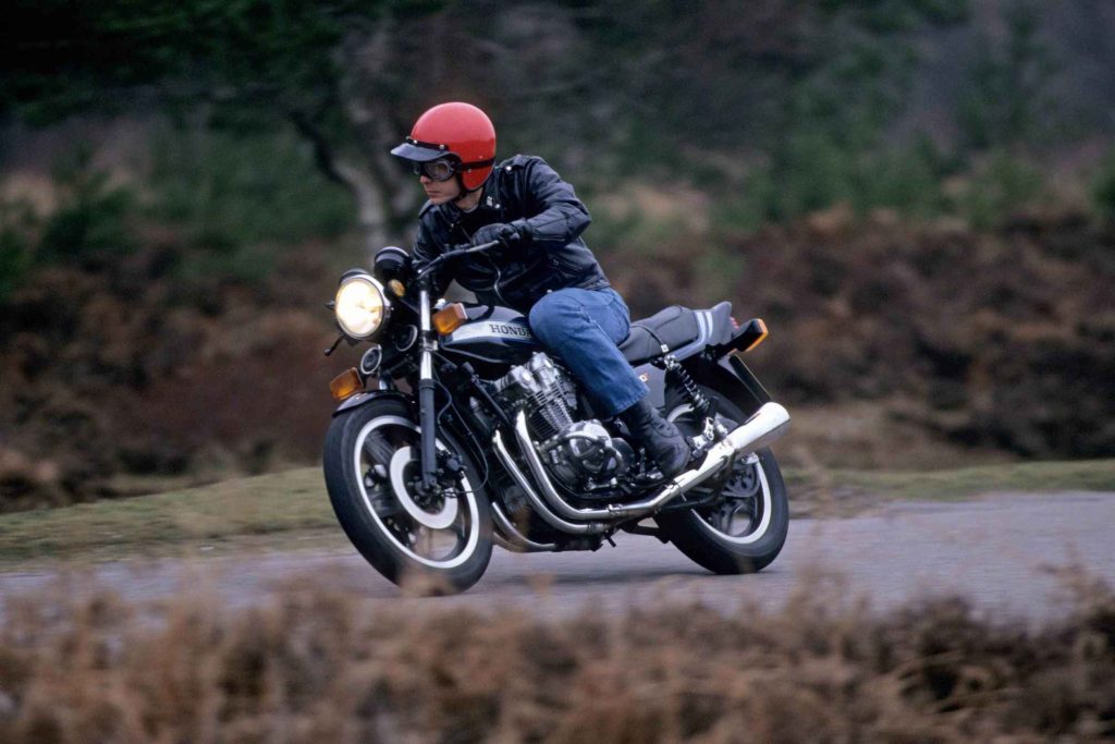 Front three-quarter action shot of a Honda CB900F being ridden on a wet road through heathland, headlight illuminated, showing the whitewall front tire and chrome exhaust pipes.
