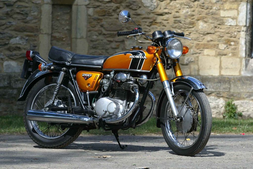 Honda CB250 K4 in gold, parked against a stone wall in bright sunshine, showing the twin-cylinder engine, spoke wheels and chrome exhaust