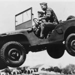 A vintage black-and-white photograph of two soldiers in uniform riding a Willys MB military Jeep mid-jump during a demonstration