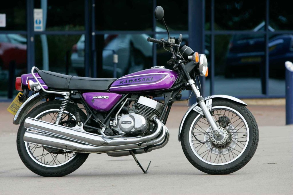Kawasaki KH400 two-stroke triple in vivid purple, parked outside a modern building, showing the three-into-three exhaust and classic 1970s styling