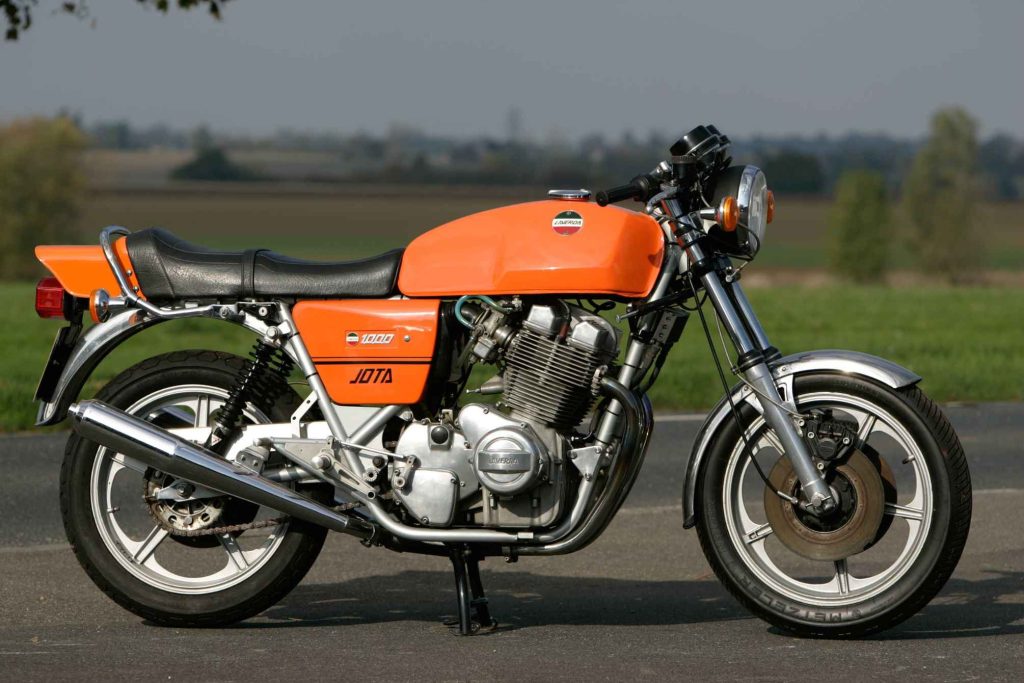 Laverda Jota 1000 in signature orange, side profile parked on tarmac with flat countryside behind, showing the triple-cylinder engine, clip-on bars and rearset footrests