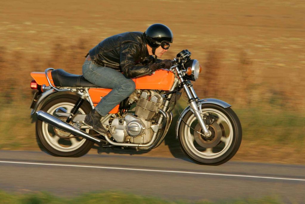 Laverda Jota in orange being ridden at speed on a country road, rider in black leather jacket and open-face helmet with goggles, late afternoon golden light