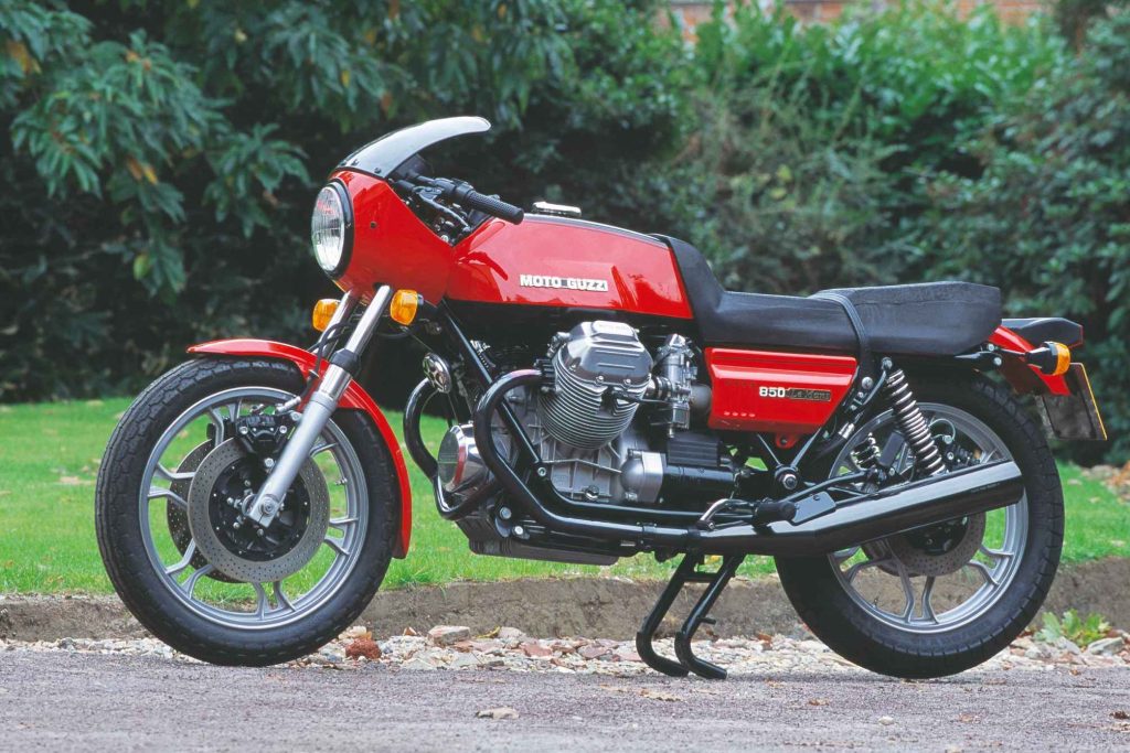 Moto Guzzi 850 Le Mans in red, parked side-on in a suburban setting with hedgerow behind, showing the transverse V-twin engine, bikini fairing and cast alloy wheels