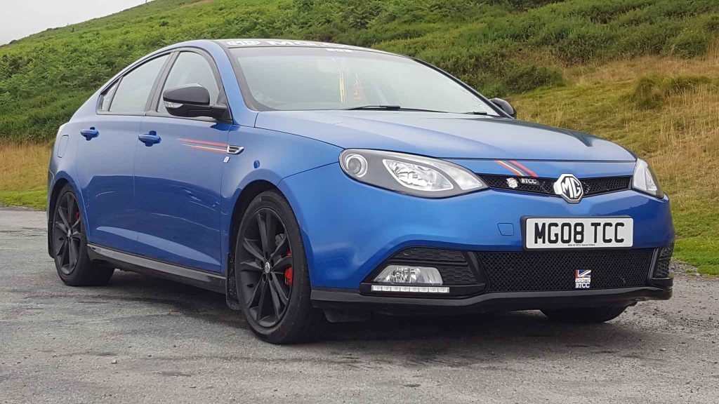 A blue MG6 BTCC Special Edition road car with black alloys, red brake calipers, and BTCC branding, parked on a road with green hills in the background, displaying the registration MG08 TCC.