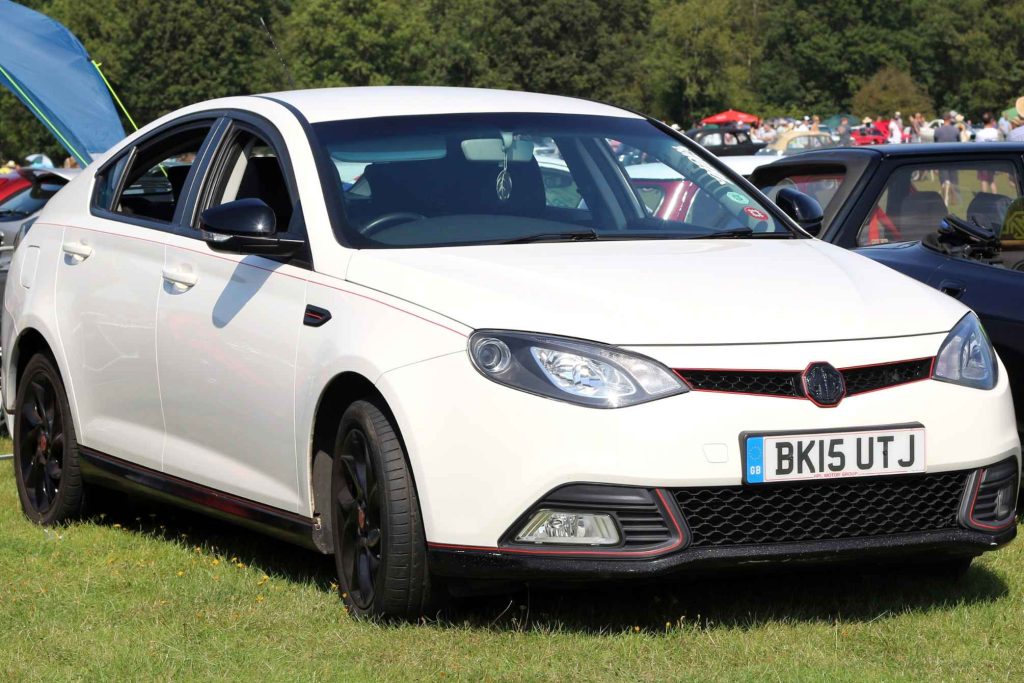 A white MG6 saloon with red accent stripes and black alloy wheels at an outdoor car show, displaying a 2015 GB registration plate (BK15 UTJ).