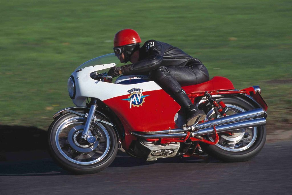 MV Agusta 750S in red, white and blue, parked side-on outside a timber building in sunshine, showing the four-cylinder engine, twin chrome exhausts and red frame