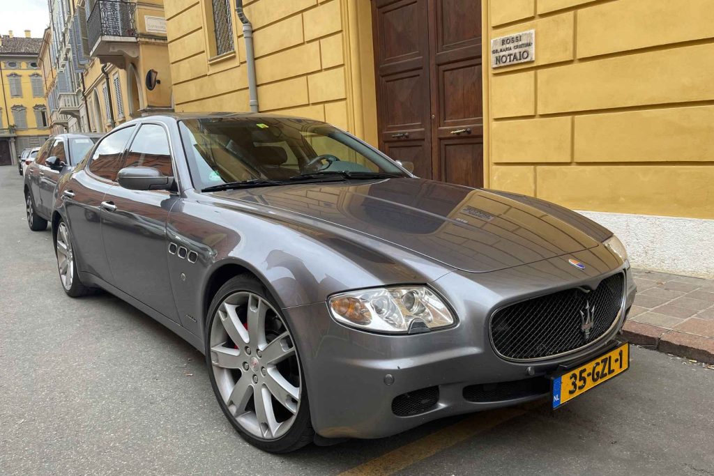 A grey Maserati Quattroporte sport sedan parked on a cobblestone street in an Italian town, with ochre-colored historic buildings behind it.