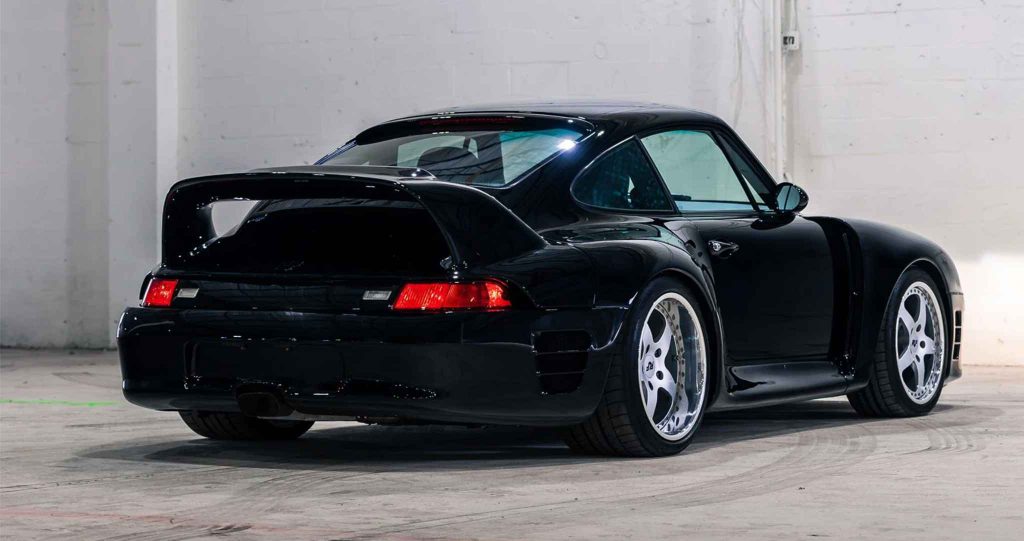 Rear three-quarter view of a gloss black Ruf CTR2 Sport based on the Porsche 993, shot in a white-walled warehouse, showing the large fixed rear wing, wide-body arches, red tail lights, and polished five-spoke RUF alloy wheels.