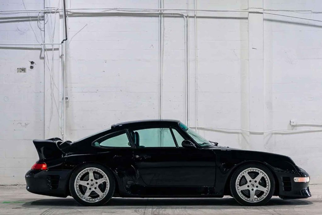 Pure side profile of the black Ruf CTR2 Sport against a white warehouse wall, clearly showing the dramatically widened bodywork, lowered stance, large rear wing, and matching polished five-spoke wheels front and rear.