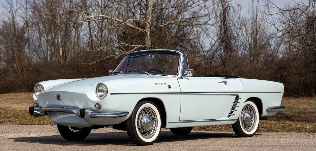 Front three-quarter view of a pale blue Renault Floride cabriolet with the roof down, showing its rounded headlights, chrome bumper, wire wheels with whitewall tyres, and decorative side vents, photographed against bare winter trees.