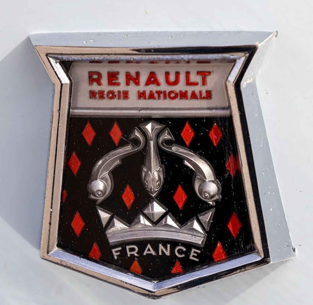 Close-up of the original Renault Régie Nationale shield badge, featuring the chrome surround, red diamond pattern on black enamel, central fleur-de-lis motif, and "France" inscription at the base.