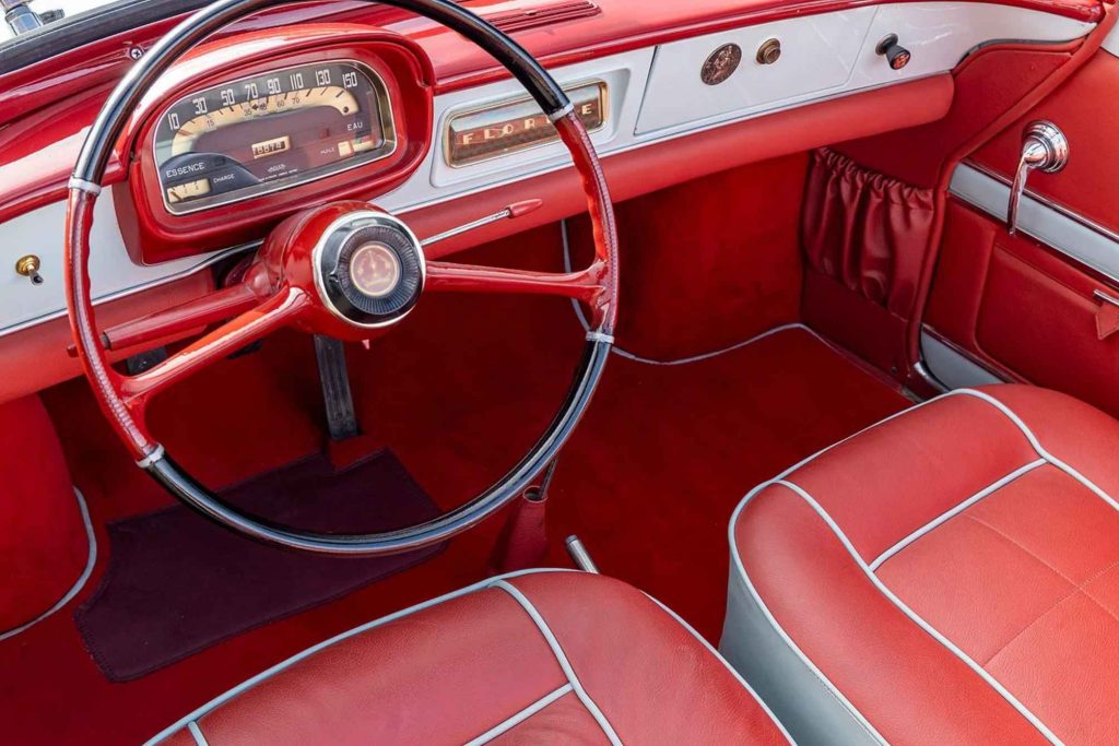 Interior of a restored Renault Floride showing the vivid red leather seats with white piping, matching red dashboard, black-rimmed steering wheel, Jaeger instrument cluster, and "Floride" script badge on the glove box.