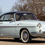 Front three-quarter view of a pale blue Renault Floride cabriolet with the roof down, showing its rounded headlights, chrome bumper, wire wheels with whitewall tyres, and decorative side vents, photographed against bare winter trees.
