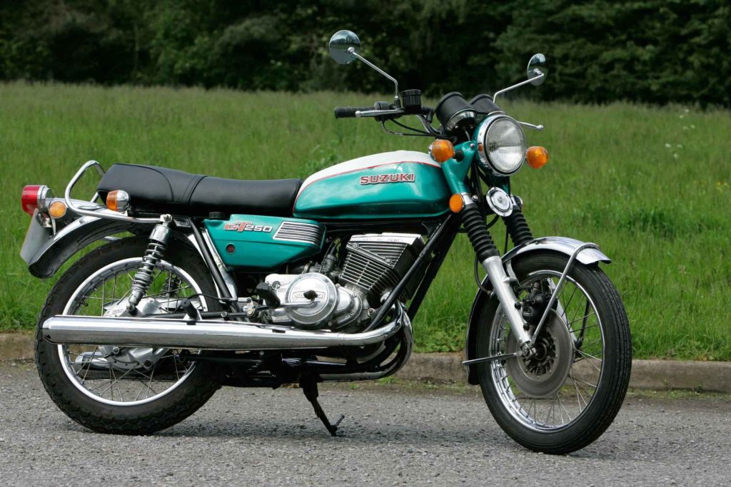 Suzuki GT250 two-stroke twin in teal green, parked side-on against a lush grass verge, showing the parallel twin engine, chrome exhausts and period-correct 1970s styling