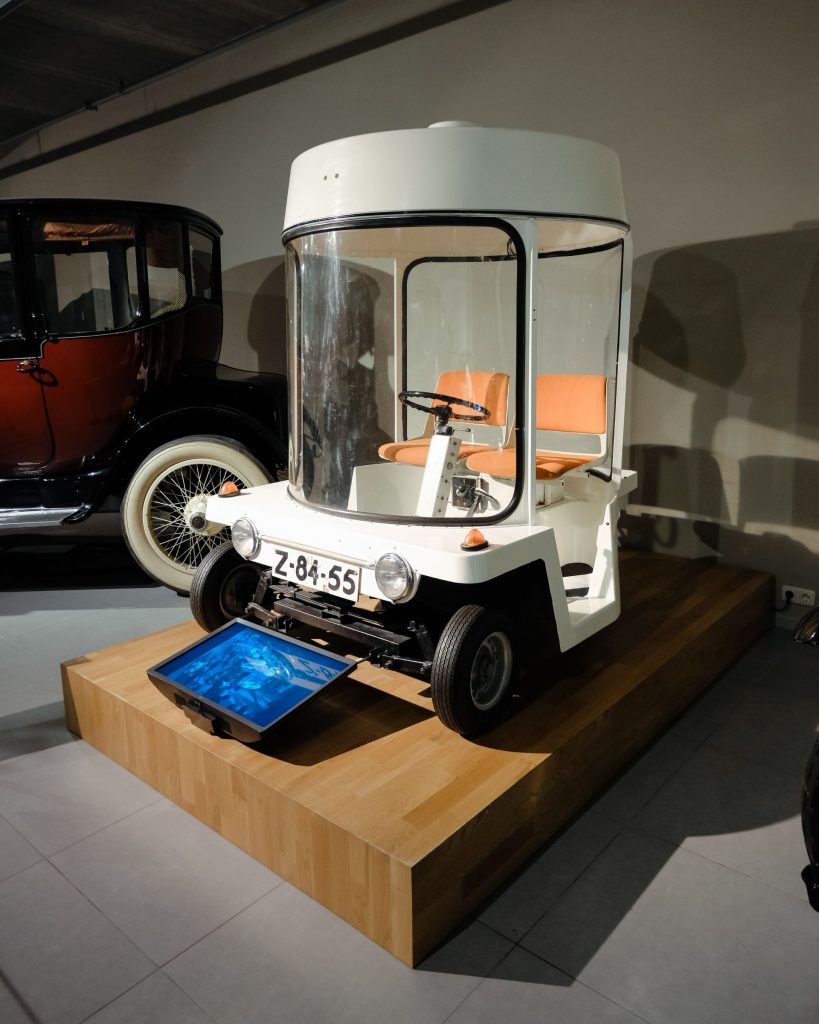 Museum display of a white Amsterdam electric city microcar on a raised wooden plinth, showing its transparent cylindrical perspex cabin, orange bench seat, simple steering wheel, and Dutch license plate "Z-84-55," with a digital screen embedded in the plinth below and a vintage black-and-red automobile visible in the background.