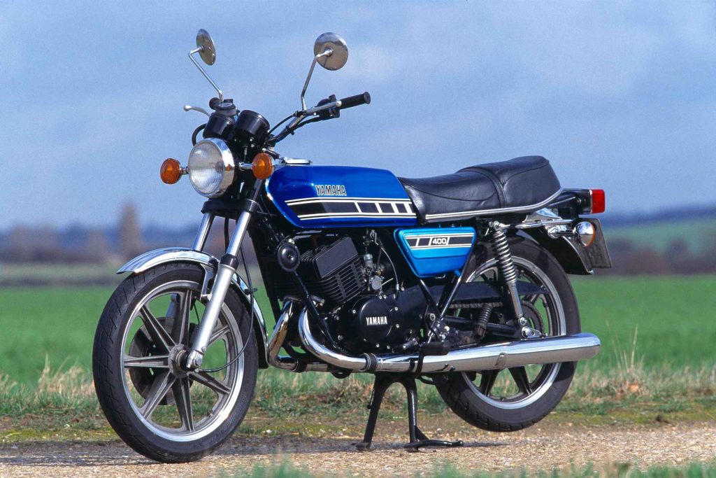 Yamaha RD400C in blue, static three-quarter front view on a rural road, showing the two-stroke parallel twin engine, cast alloy wheels and clean 1970s styling against an open sky
