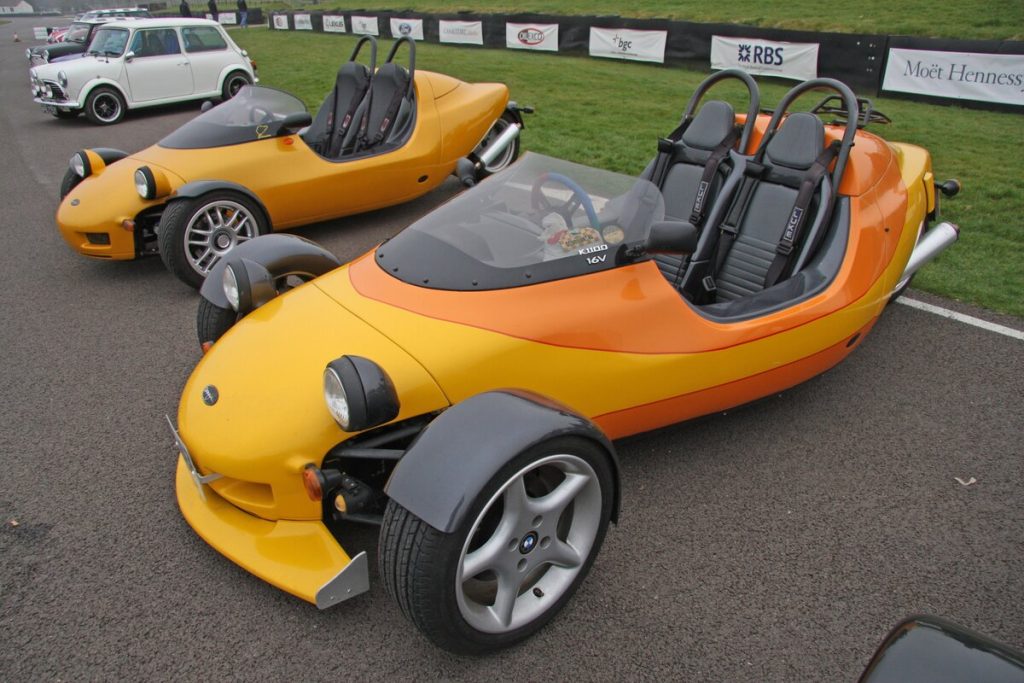 Two yellow and orange Grinnall Scorpion III BMW-powered three-wheelers parked in the pit lane at Goodwood Circuit