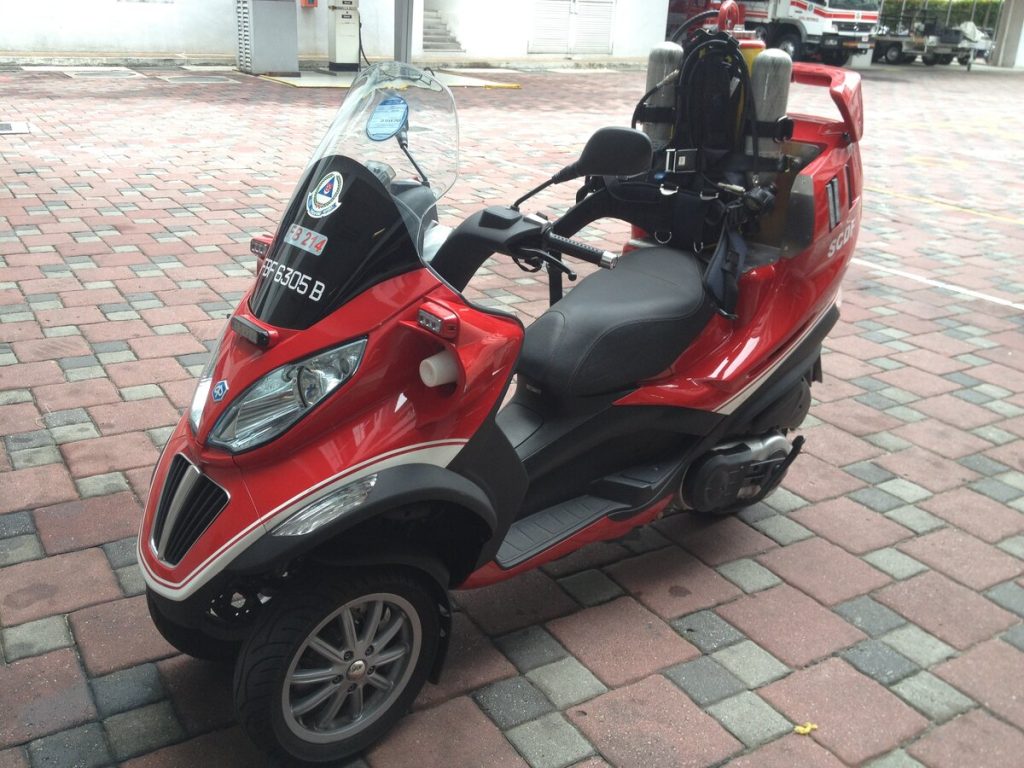 Red Singapore Civil Defence Force (SCDF) Piaggio MP3 three-wheel emergency response scooter parked outside a fire station on brick pavers.