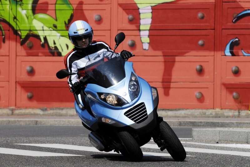 A rider in a white and red motorcycle jacket and blue helmet cornering a light blue Piaggio MP3 three-wheel tilting scooter
