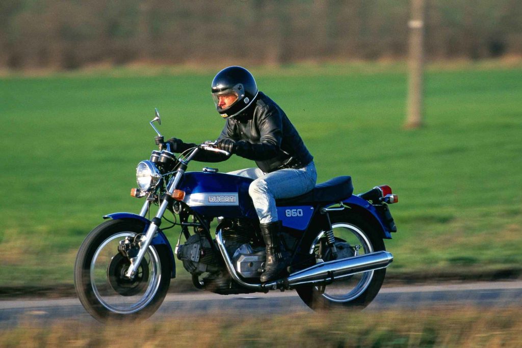 Rider in black helmet and leather jacket riding a classic Ducati 860 on a country road