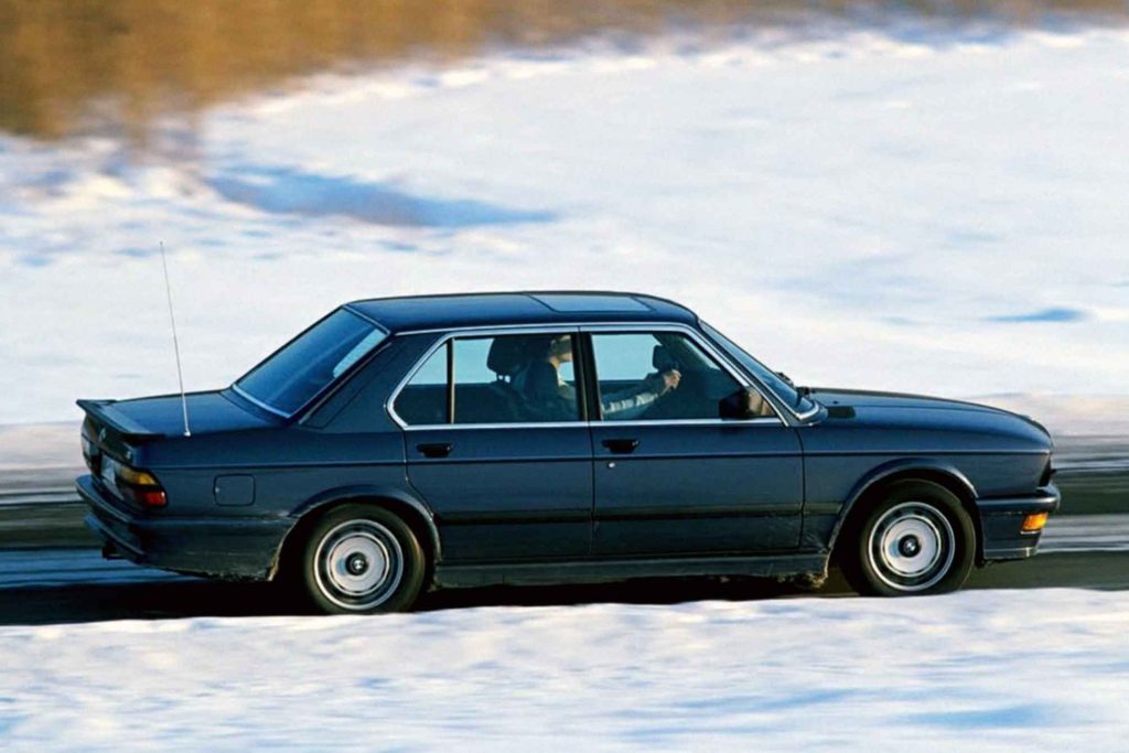 Dark blue BMW M535i E28 five-series saloon in left-side profile, being driven at speed along a snowy winter road