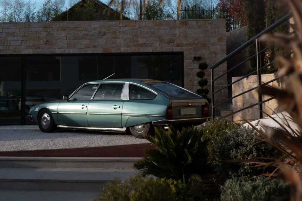 Left-side profile of a light metallic green Citroën CX parked on a gravel drive outside a contemporary stone-clad villa at dusk