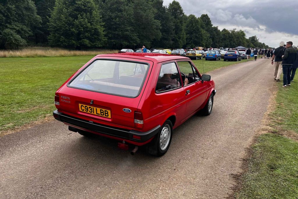 Rear three-quarter view of a red Ford Fiesta Mk2 Popular three-door hatchback being driven along a gravel path