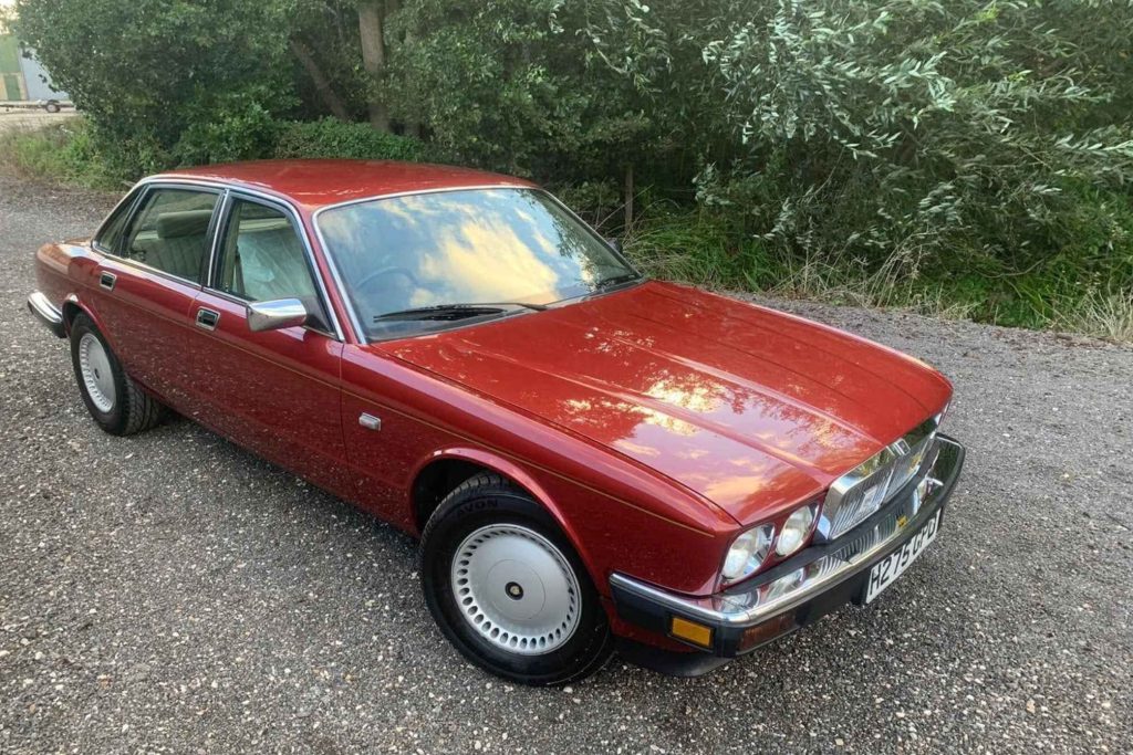 Front three-quarter view of a metallic red Jaguar XJ6 Series III four-door saloon parked on gravel in front of trees
