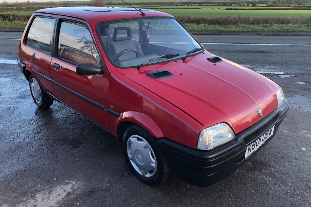 Front three-quarter view of a red Rover Metro three-door hatchback parked on a wet road with open farmland behind.