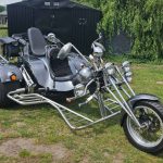 Custom-built chrome and black chopper-style three-wheel trike with extended front forks parked on grass in a rural setting.